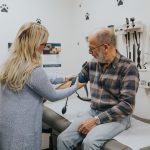 A female physician takes the blood pressure of a male patient as he sits on the bed of a physician's office.