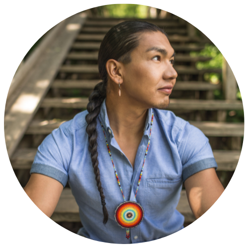 Biography picture of Dr. Makokis. He is sitting on a staircase in the outdoors looking off to the right with a pensive look on his face. He is wearing a blue shirt with an Indigenous beaded necklace and his hair is in a braid.