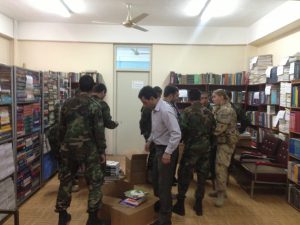 A group of people standing in the centre of a room. Among the people are members of the Canadian military. There are bookshelves lining the walls and the people are unloading medical books from boxes.