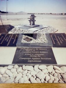 A monument with a number of plaques sits on the ground outside. A stone Inukshuk sits on top of the plaques. The monument is outside in a desert-like environment with hills in the background.