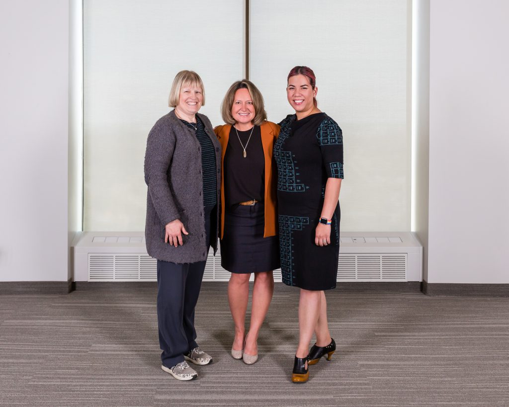 Three women posing for a picture in front of a window. The woman on the left has short blonde hair and is wearing a grey sweater and dark pants. The woman in the middle has short brown hair and is wearing a dark dress with an orange cardigan and the woman on the right has her dark red hair pulled back into a ponytail and is wearing a black and blue dress.