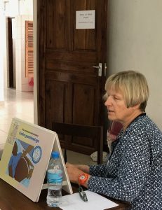 A woman with short blonde hair and a patterned blue blouse sits at a table looking at a teaching resource that is propped up on the table. There is a plastic water bottle on the table and the door to the room is open.