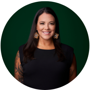 A woman wearing a black top and flower earrings smiling for a photo against a dark green backdrop.