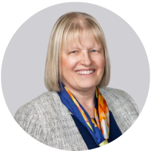 A woman with short blonde hair wearing a grey blazer and a orange and blue scarf smiles for a picture.