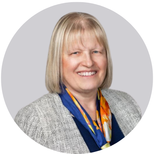 A woman with short blonde hair wearing a grey blazer and a orange and blue scarf smiles for a picture.