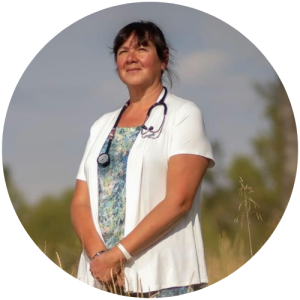 A woman with brown hair standing in a field wearing a white doctor's coat and a stethoscope around her neck.