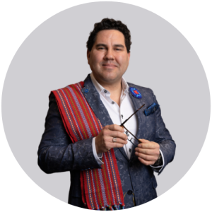 A man with dark brown hair smiling for a professional headshot. He is wearing a dark blue/grey suit with a Metis sash over his right shoulder and is holding his glasses in his hands.